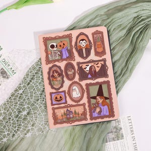 May include: A light pink tablet case featuring framed Halloween-themed illustrations. The illustrations include a skeleton, pumpkin, vampire, witch, ghost, and haunted house. The case is decorated with brown frames and a black border.