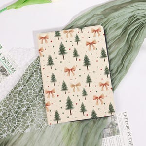 May include: A notebook with a cream-colored cover featuring a pattern of green Christmas trees, red bows, and small red dots. The notebook has a black spine and is set against a green and white background.