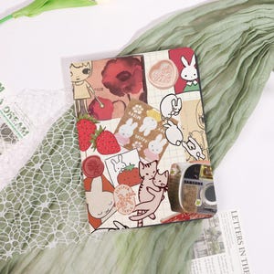 May include: A decorative journal with a collage of illustrations, including strawberries, bunnies, cats, and a vintage camera. The cover features a variety of stickers and stamps in shades of red, brown, and beige, with a green fabric in the background.