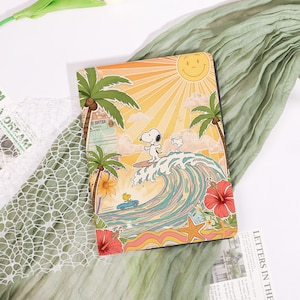 Peut inclure: Un carnet illustré avec Snoopy surfant une vague sur une plage ensoleillée. Le design comprend des palmiers, un soleil souriant et une voiture classique. Le carnet est rectangulaire et présente une couverture illustrée colorée.