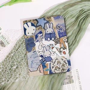 May include: A decorative journal featuring a collage of illustrations, including a blue bunny, a girl with an umbrella, and a cat. The journal has a blue and beige colour scheme with Japanese text and a circular sticker that says "The Good, The Bad, The Unique."