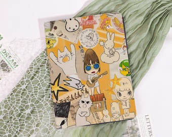 Könnte beinhalten: Ein beigefarbener und gelber Notizbuchumschlag mit Cartoon-Illustrationen von Katzen, einem Mädchen, das Gitarre spielt, und dem Text "LOLITA BANANAS". Umgeben von einem grünen Schal und weißem Spitzengewebe.