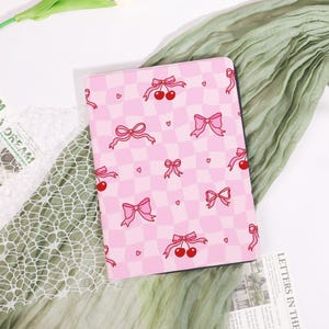 May include: A pink tablet case with a checkered pattern featuring pink bows, cherries, and hearts. The case is designed with a playful aesthetic, perfect for protecting a tablet. The case is made of a durable material.