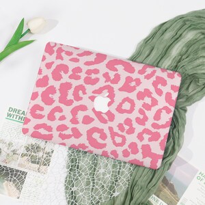 May include: Pink and light pink leopard print laptop case with the Apple logo. The case is on a white surface with a green scarf and a white tulip. The text "DREAM WITHI" is visible.