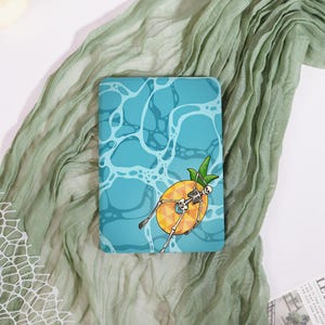 May include: A rectangular e-reader case featuring a graphic of a skeleton floating on a yellow pineapple-shaped pool float. The background depicts a light blue swimming pool with white water ripples and light reflections across the surface.