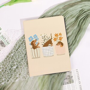 Peut inclure: Un cahier beige avec une illustration de trois chiens teckel dans des pots de fleurs. Les chiens sont bruns, avec différentes fleurs dans chaque pot. Les pots ont des motifs rayés, à carreaux et rayés.