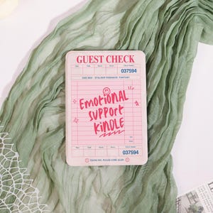 May include: A pink guest check with the words "GUEST CHECK" at the top and "EMOTIONAL SUPPORT KINDLE" in the center. The check also includes the text "ONE BED - STALKER ROMANCE - FANTASY" and the number "037594".