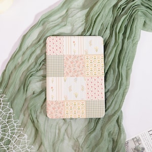 May include: A rectangular notebook with a patchwork design featuring floral and gingham patterns in soft pink, cream, and green hues. The notebook is set against a backdrop of a flowing, sage green fabric.