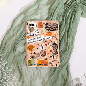 May include: A rectangular phone case with a fall-themed design. The case features a ghost reading a book, pumpkins, a skeleton, and the text "Another fall, another page turned..." and "Fall is my favorite color". The color palette includes orange, brown, and cream.
