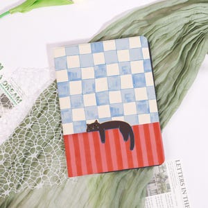 May include: A rectangular notebook with a whimsical design. The top half features a blue and white checkered pattern, while the bottom half has red and pink stripes. A black cat illustration is resting on the stripes. The notebook is on a white surface.