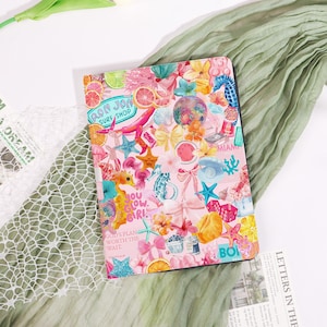 May include: A pink notebook cover with a colorful collage of beach-themed illustrations, including starfish, seahorses, and flowers. The cover features text that reads "RON JON SURF SHOP", "YOU GLOW GIRL", and "GOD'S PLAN WORTH THE WAIT".