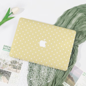 May include: A yellow laptop case with a white polka dot design. The case has a white Apple logo. The laptop case is on a white surface with green fabric and a white flower.
