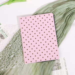May include: A pink notebook with a brown polka dot pattern. The notebook is rectangular and has a black border. The background includes a green fabric and white lace.