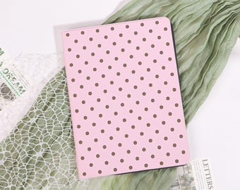 Op de afbeelding: Een roze notitieboek met een bruin stippenpatroon. Het notitieboek is rechthoekig en heeft een zwarte rand. De achtergrond omvat groene stof en wit kant.