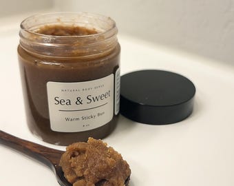 May include: A jar of "Sea & Sweet" body scrub with a "Warm Sticky Bun" scent, next to a wooden spoon holding a scoop of the scrub. The jar has a black lid and a white label. The scrub is brown.