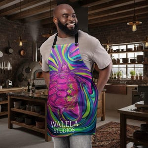 May include: A colourful apron with a striking turtle design and swirling patterns in pink, purple, blue, and green. The apron has black straps and the text "WALELE STUDIOS". This kitchen accessory is ideal for cooking and baking.