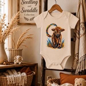 Peut inclure: Un body pour bébé à manches courtes de couleur crème, orné d'une illustration graphique d'une vache highland brune debout dans de l'herbe bleue et jaune avec un accent circulaire aux couleurs de l'arc-en-ciel derrière l'animal, sur un cintre en bois.