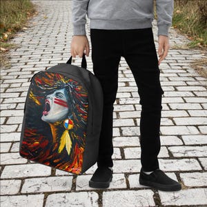 May include: Black backpack featuring a vibrant painting of a Native American woman with red face paint and an open mouth. The artwork includes colorful hair and a feather earring. The backpack is held by a person.