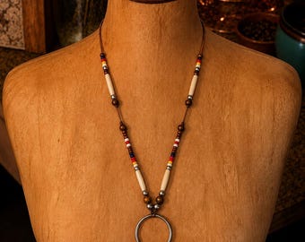 Puede incluir: Un collar con un cordón marrón ensartado con cuentas blancas, rojas, amarillas, azules y plateadas metálicas. El diseño incluye cuentas cilíndricas blancas largas y un anillo central de metal plateado con cuatro flecos de cuentas colgantes.