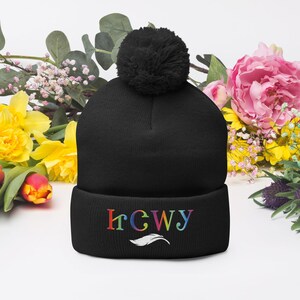 Op de afbeelding: Zwarte gebreide muts met een grote pompon. De rand heeft de geborduurde tekst "IrGWy" in regenboogkleuren met een wit veermotief eronder. De muts wordt omringd door kleurrijke bloemen.