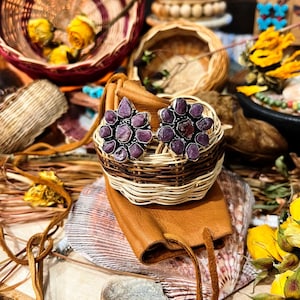 May include: A pair of stud earrings featuring clusters of raw, deep purple ruby stones set in silver-toned metal. Each earring is shaped like a flower with multiple irregular, faceted stone petals surrounding a central stone, creating a textured look.