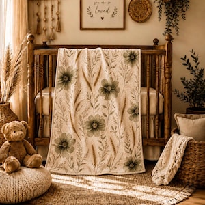 May include: A cream-colored baby blanket featuring a botanical print of sage green flowers and golden wheat stalks. The rectangular blanket is draped over a wooden crib, showcasing a repeating floral pattern with thin stems and leaves across the fabric.