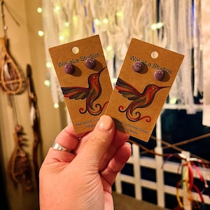May include: Two pairs of round purple stud earrings mounted on brown cardboard display cards. Each card features the text Wal aLa Studios and a colorful hummingbird illustration, with the text Raw Natural Turquoise Stones Indigenous Made.