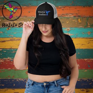 May include: Black and white trucker hat with the text "WaLeLa Studios" and a hummingbird logo. The hat has a black brim and crown, with a white mesh back. The hummingbird logo is in shades of blue, pink, and yellow.