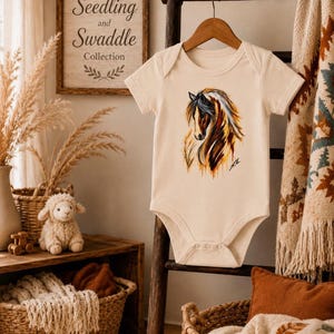 May include: A cream-colored short-sleeve baby bodysuit featuring a detailed, artistic illustration of a horse head with flowing mane in shades of brown, black, and white. The garment has a round neckline and snap closures at the bottom crotch area.