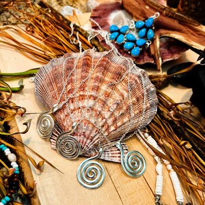 May include: Four silver-toned spiral pendants on thin metal chains resting on a large, fan-shaped brown and white seashell. A separate cluster of bright blue teardrop-shaped stones is visible nearby, along with white disc-shaped beads on a string.