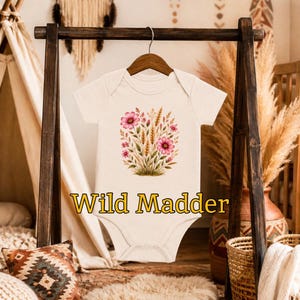 May include: A cream-colored short-sleeve baby bodysuit featuring a central graphic of pink wildflowers and wheat stalks. The text "Wild Madder" is printed in yellow letters below the floral illustration on the front of the garment.