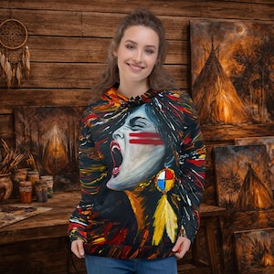 Puede incluir: Una sudadera con capucha negra con un retrato impactante de una mujer nativa americana con pintura facial roja, la boca abierta y un pendiente de plumas de colores. El diseño incorpora trazos audaces de rojo, amarillo y azul. El fondo es una pared de madera con pinturas.