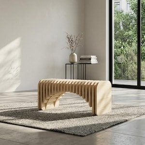 Könnte beinhalten: Eine hellfarbene Holzbank mit geschwungenem Lamellendesign. Die Bank steht auf einem grau strukturierten Teppich in einem hellen Raum. Eine Vase mit Zweigen und Büchern ist im Hintergrund zu sehen.