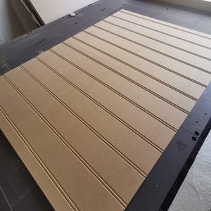 May include: A large, rectangular sheet of light brown medium-density fiberboard featuring evenly spaced, vertical grooves cut into the surface. The board is positioned on a dark industrial machine bed with a smooth, matte finish.