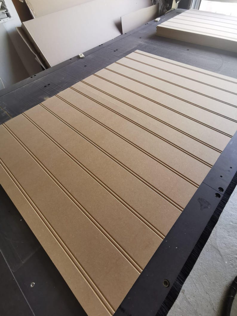 May include: A large, rectangular sheet of light brown medium-density fiberboard featuring evenly spaced, vertical grooves cut into the surface. The board is positioned on a dark industrial machine bed with a smooth, matte finish.