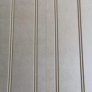 May include: A light gray panel featuring vertical beadboard detailing. The surface has a smooth, matte texture with evenly spaced, thin, recessed vertical grooves running parallel from the top to the bottom edge of the rectangular board.