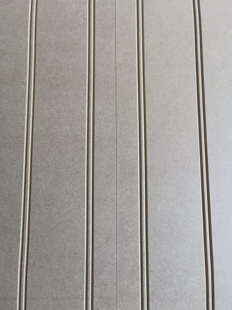 May include: A light gray panel featuring vertical beadboard detailing. The surface has a smooth, matte texture with evenly spaced, thin, recessed vertical grooves running parallel from the top to the bottom edge of the rectangular board.