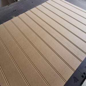 May include: A large, rectangular sheet of light brown medium-density fiberboard featuring evenly spaced, vertical grooves running across its surface. The board is positioned on a dark, industrial-style work surface with visible mounting points.