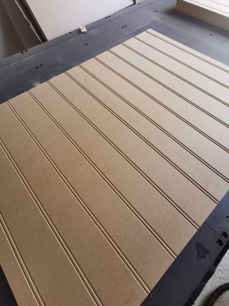May include: A large, rectangular sheet of light brown medium-density fiberboard featuring evenly spaced, vertical grooves running across its surface. The board is positioned on a dark, industrial-style work surface with visible mounting points.