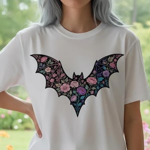 May include: White t-shirt featuring a floral bat design. The bat is black with a detailed floral pattern in shades of pink, purple, and blue. The shirt has short sleeves and a crew neck. The design is centered on the front of the shirt.