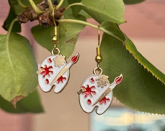 May include: A pair of gold-colored dangle earrings. Each earring features a white enamel artist's palette design with red paint splotches and a paintbrush. The earrings are hanging from gold-colored hooks.