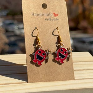 Puede incluir: Un par de pendientes hechos a mano con un diseño de tiburón de dibujos animados rojo y ganchos dorados. Los pendientes se exhiben en una tarjeta marrón con el texto "Handmade with love."