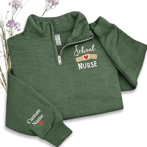 May include: A green quarter-zip sweatshirt with the words "School Nurse" embroidered on the chest, along with a bandage and heart design. The left sleeve has "Custom Name" embroidered with a red lip graphic. The sweatshirt is made of a soft material.
