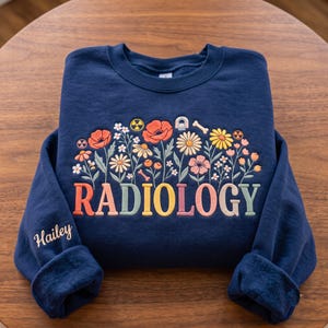 May include: A navy blue crewneck sweatshirt featuring an embroidered floral design with the word RADIOLOGY in multicolored letters. The design includes small icons of a bone, an MRI machine, and radiation symbols. The name Hailey is on the sleeve.