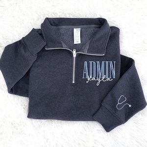 May include: Dark gray quarter-zip sweatshirt with the word "ADMIN" and the name "kayla" in light blue script. A stethoscope graphic is embroidered on the sleeve. The sweatshirt has a silver zipper and a folded collar.