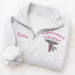 May include: A light gray quarter-zip sweatshirt with the name "Rosie" embroidered in pink on the left chest. The right chest features the text "certified surgical technologist" and a medical symbol in pink and black.