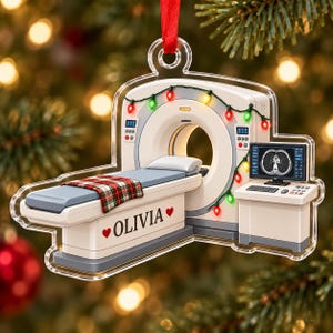 May include: A clear acrylic Christmas ornament shaped like a CT scanner machine. The scanner is decorated with colorful string lights and features a plaid blanket on the bed. The name OLIVIA is printed on the side between two red hearts.
