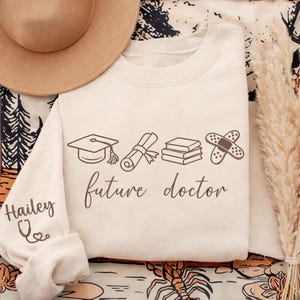 May include: Cream-colored sweatshirt with the text "future doctor" and medical-themed graphics. The sleeve has the name "Hailey" and a stethoscope design. The design includes a graduation cap, diploma, books, and a bandage.