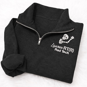 May include: A dark gray quarter-zip sweatshirt with a silver zipper. The sweatshirt features a white embroidered skeleton graphic and the text "Lauren RT(R) Rad Tech". The sleeves are folded.