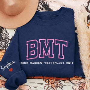 May include: Navy blue sweatshirt with the letters "BMT" in pink, outlined in a collegiate style. The words "Bone Marrow Transplant Unit" are below the letters. The sleeve has the name "Sophia" and a heart.